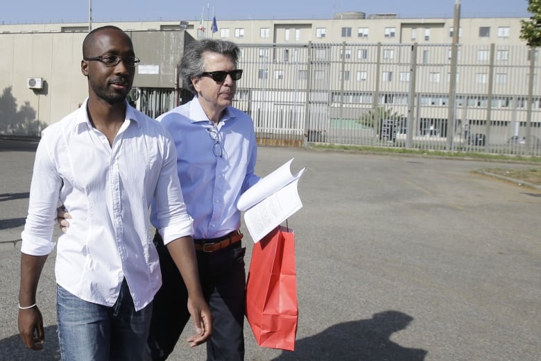 A dark skinned man with some facial hair and glasses is shown walking outdoors wearing jeans and a white collared shirt.