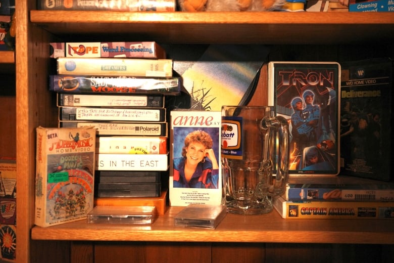 A bookcase holds a number of video tapes. One is an Ann Murray concert film. Another is the VHS for Tron. Next to them is a stack of tapes, many of them homemade. 