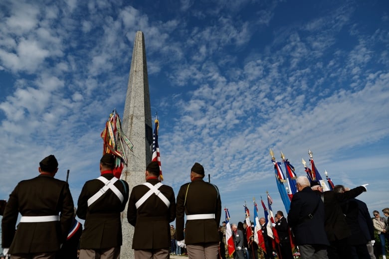 A group of people wearing military regalia stand at a long pole-shaped memorial on a cloudy day.