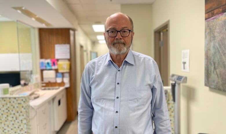 Dr. Rick Ward, has a beard and glasses. He's standing in a clinic hallway. Medical supplies and a scale can be seen behind him.
