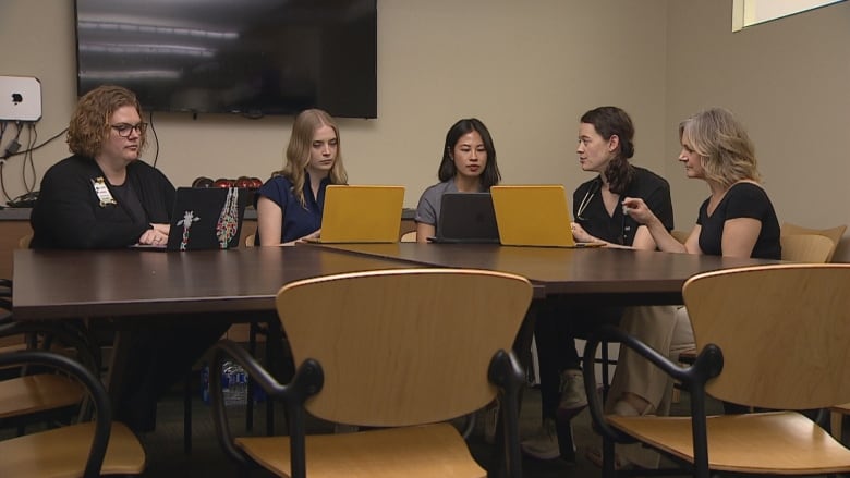 Five women gather at a conference room table. They're looking at laptops