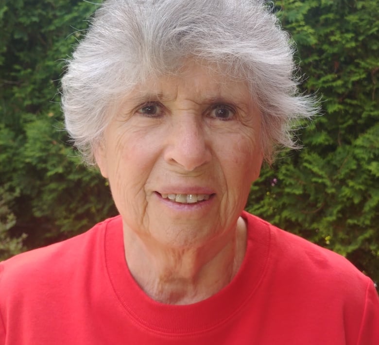 A woman stands outside, looking to camera. She is wearing a pink top, standing in front of a hedge.