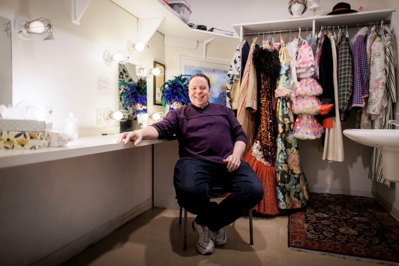 A man sits in a dressing room, smiling. 