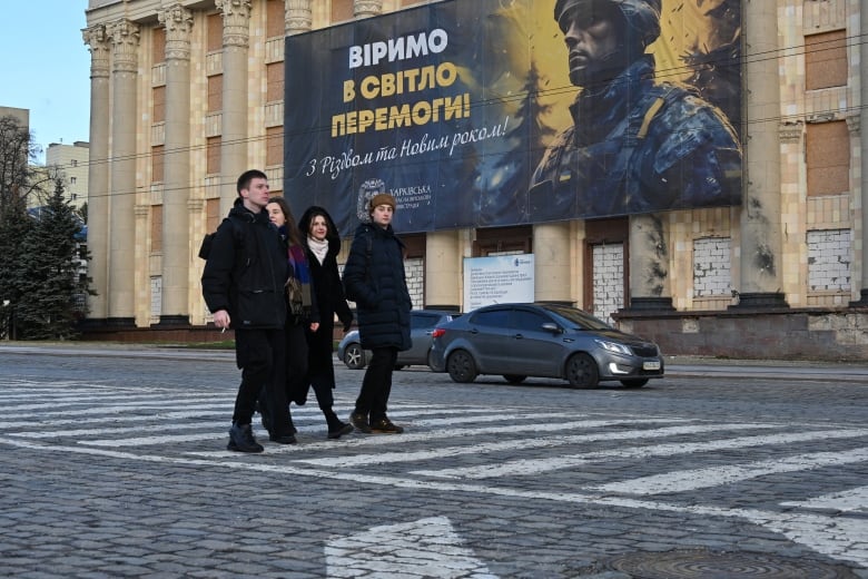 Four people walk in a crosswalk on a city street. A large billboard on the building behind them displays a soldier next to a message with Cyrillic letters.