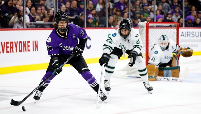 A female ice hockey player controls the puck as a defender reaches at them with their stick during a game as fans watch from behind the glass.