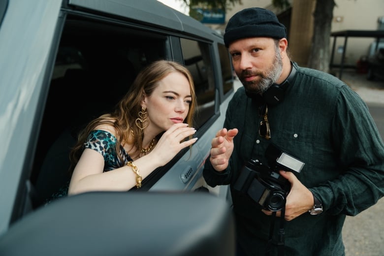 Emma Stone talks to director Yorgos Lanthimos on the set of Kinds of Kindness. She is sitting in a car and cupping her hand near her mouth.