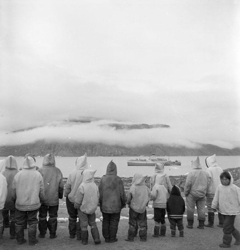 A black and white photo. A group of Inuit stand and look at an incoming ship.