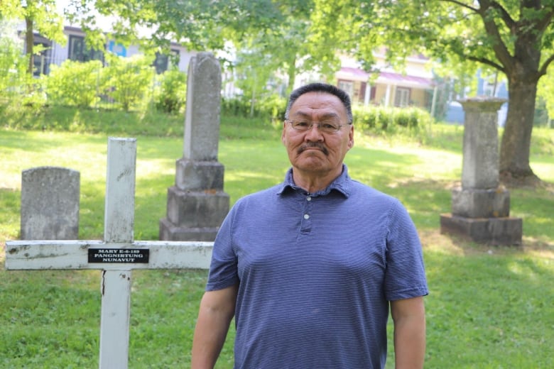 A man in a blue shirt looks at the camera. He is standing in a graveyard, next to a white cross. 