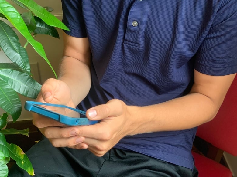 A young man's torso and hands are seen as he scrolls his phone while sitting on a bright pink chair next to a houseplant.