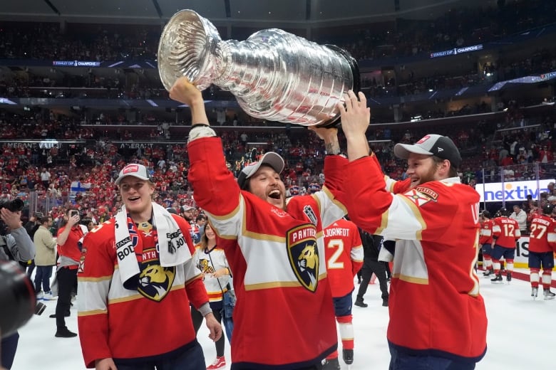 Flanked by two other players, a hockey player in a baseball cap lifts a huge silver trophy over their head. 