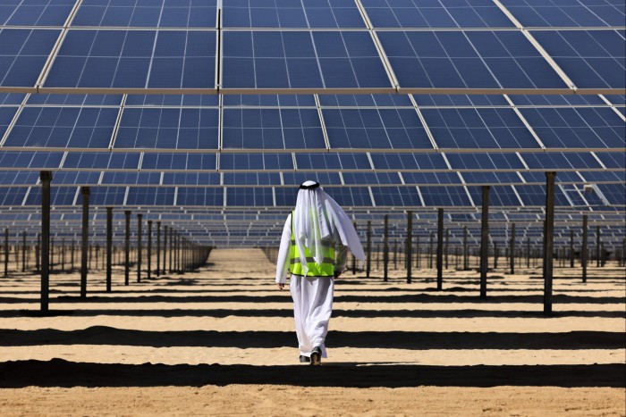 A man in traditional Middle Eastern attire and a reflective safety vest walks through a solar farm with large solar panels arranged in rows