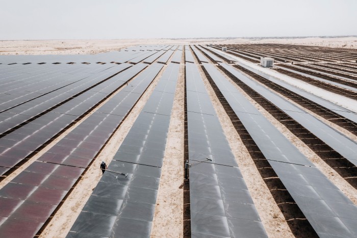 Aerial view of a large-scale solar energy facility in a sandy desert, showing numerous rows of solar panels and two maintenance workers in the middle