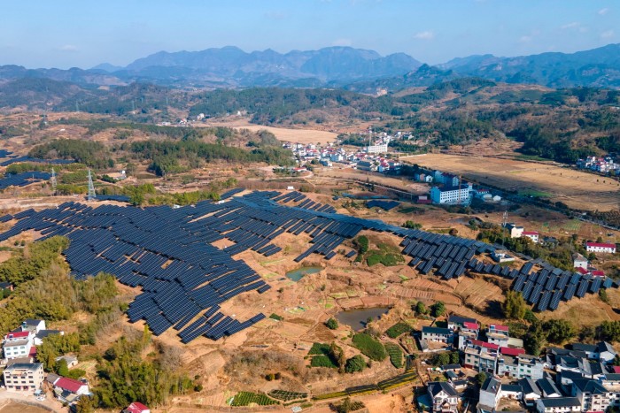 Aerial view of a large solar farm situated in a rural landscape, with rows of solar panels spread across the fields, surrounded by trees, small ponds, and nearby villages with mountainous terrain in the background