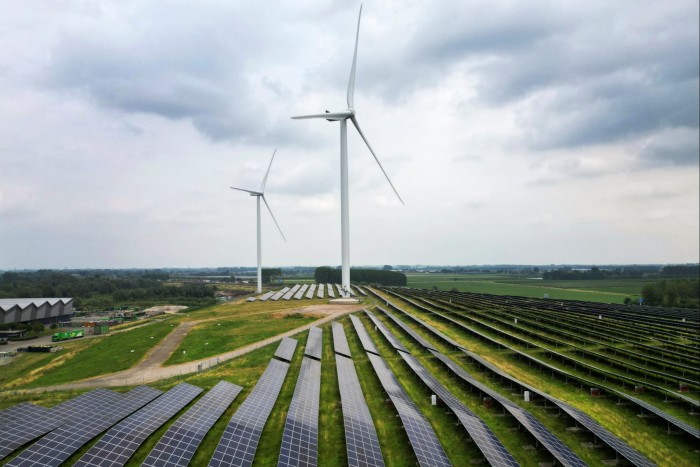 A renewable energy farm featuring rows of solar panels and several wind turbines standing tall in a green field under a cloudy sky