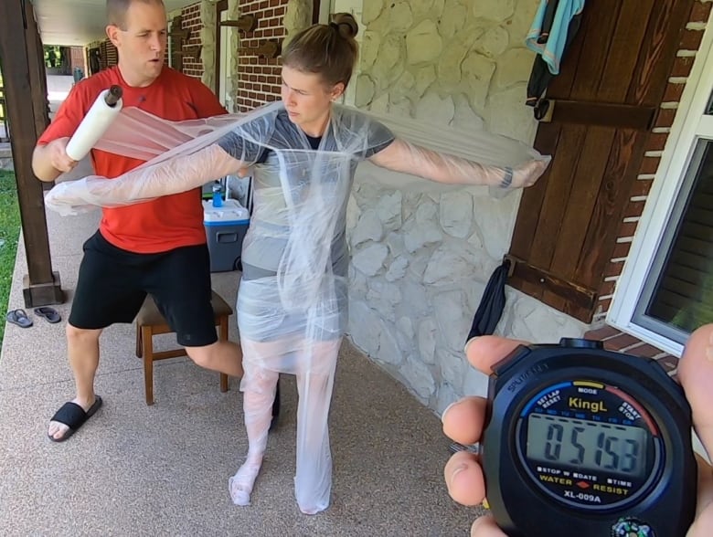 A man stands on a porch wrapping a woman in cling wrap