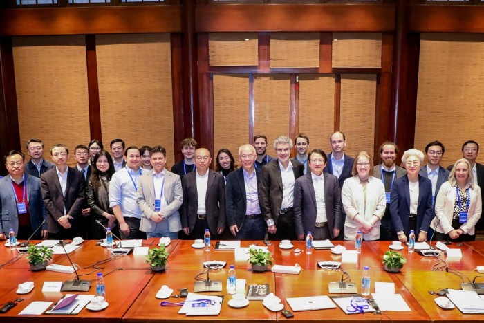 A diverse group of professionals stands together for a photo in a well-lit conference room. The long table in front of them is equipped with water bottles, papers, and microphones