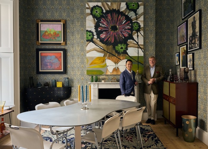 The couple stand behind a kidney-shaped table with modern paintings behind on a wall with period wallpaper