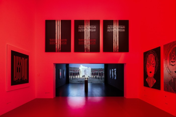 An art gallery space, lit in red, with artworks on the wall. Just visible in the next gallery is a portrait of Putin in flames and flanked by Pussy Riot members in their trademark balaclavas