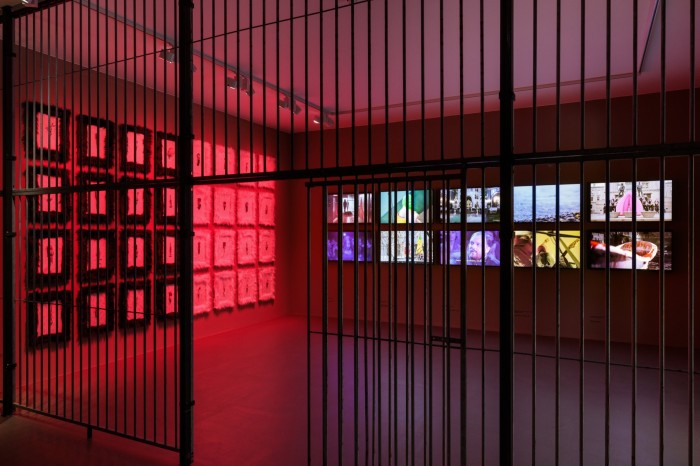 A gallery space behind bars and lit in red, with a bank of TV screens on the far wall