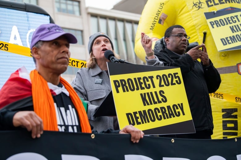 A man holds a yellow and black protest sign reading "Project 2025 KILLS DEMOCRACY" next to two other protesters. 