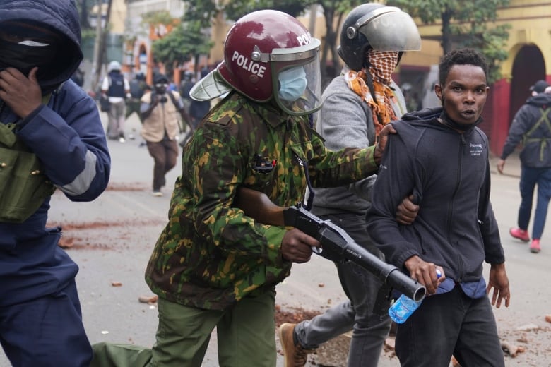 A man in the streets being led away by the arm by two armed police officers in riot helmets