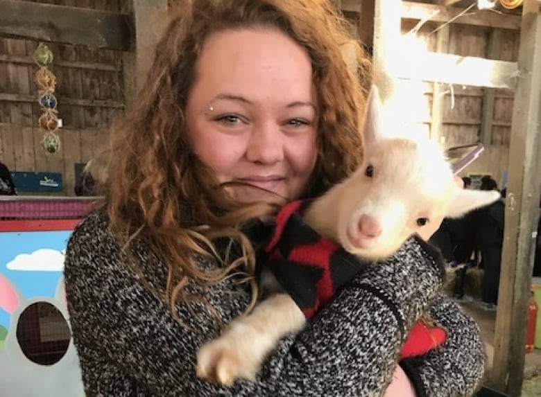 A young woman with long brown hair cuddles a small white goat. 