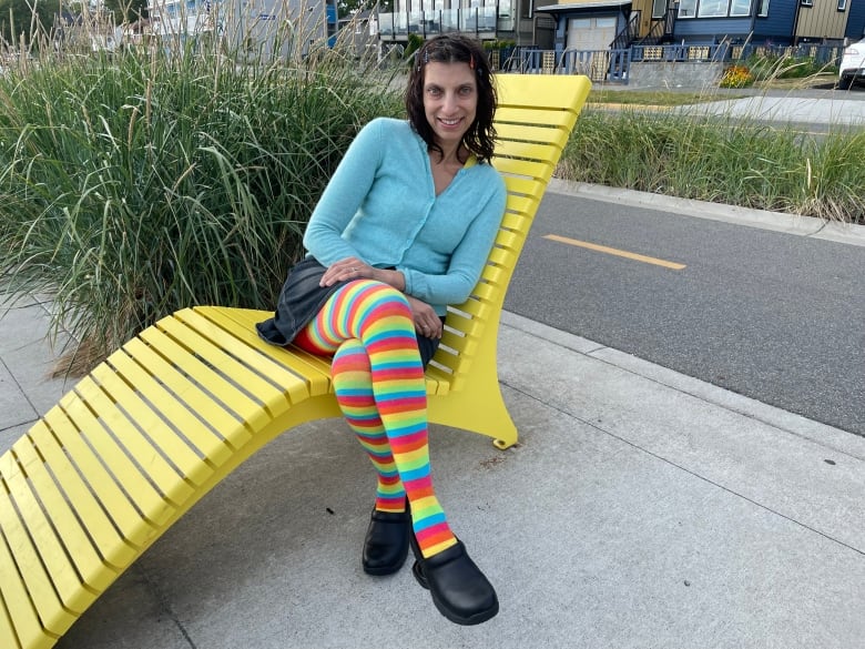 A smiling woman wearing multicoloured tights and a blue sweater sits on a yellow outdoor chaise.