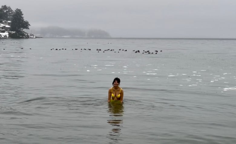 A smiling woman in a yellow bikini stands half-submerged in a grey-coloured water body.  