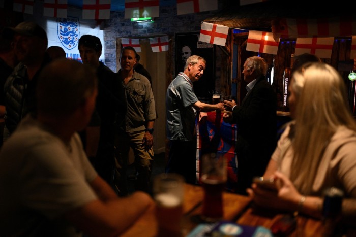 A man in a blue football shirt,  holding a pint of beer, stands talking to a man in a pub where  red-and-white St George’s Cross flags decorate the bar