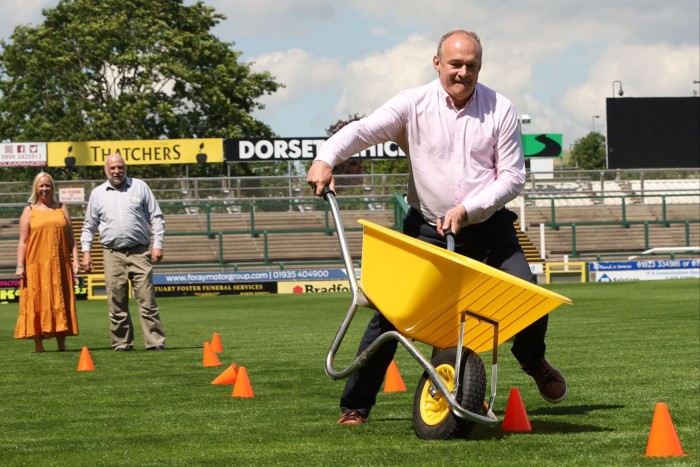 A middle-aged man in suit trousers and pink shirt pushes a yellow wheelbarrow around cones set out on a football pitch as a man and a woman stand behind him smiling