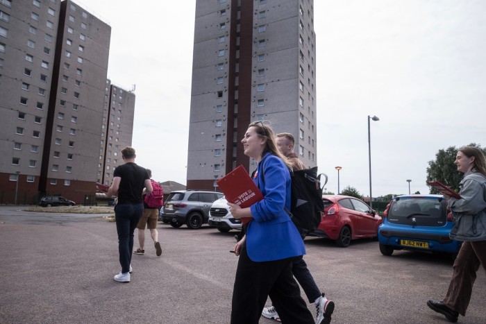 A smiling young woman in a blue jacket strides with four other people near a high rise apartment block. Like the others, she carries a red clipboard with the words VOTE LABOUR