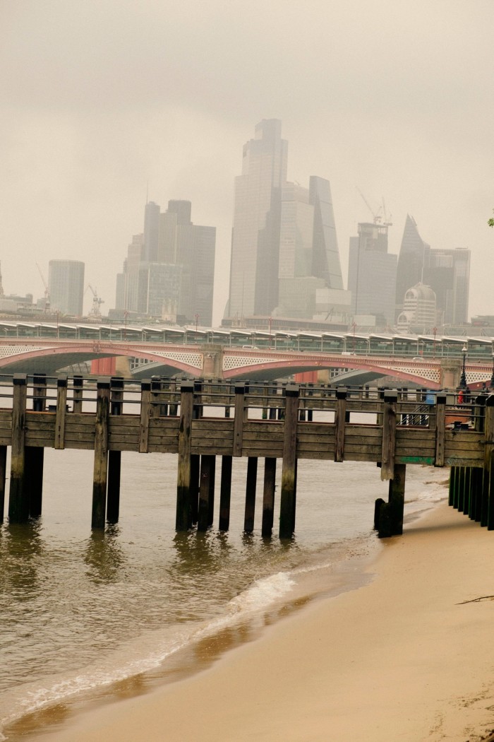 A view from a Thames river bank towards a cluster of skyscrapers shrouded in mist