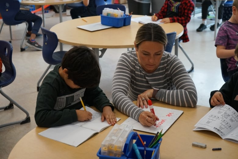 A student writes in a notebook while a teacher helps.