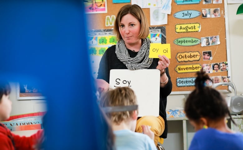 A teacher sits in front of her young class, holding up two small cards marked 'oy' and 'oi' in her left hand, with the word 'soy' written on a whiteboard balanced on her lap.