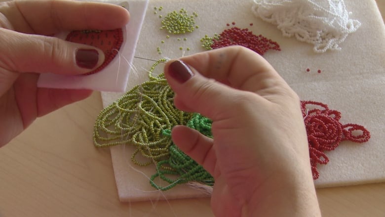 A close-up of hands working on a beaded image of a strawberry, with green, red, white beads seen on the table underneath. 