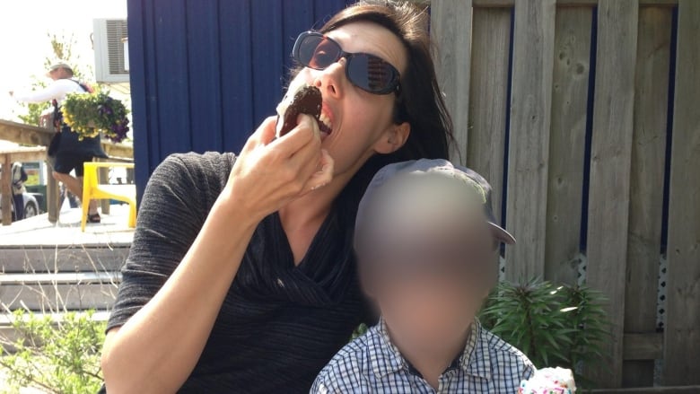 A woman eats an ice cream sandwich while sitting next to a boy whose face has been blurred. He’s holding a half-eaten ice cream cone with rainbow sprinkles. 