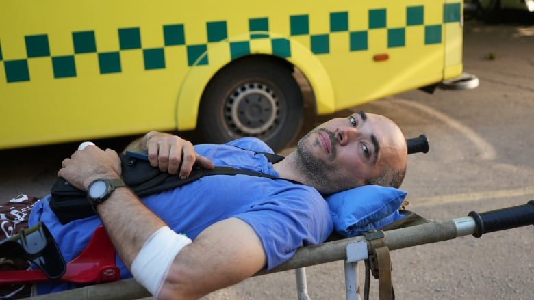 A man in a blue shirt lays on a stretcher in front of a yellow ambulance. 