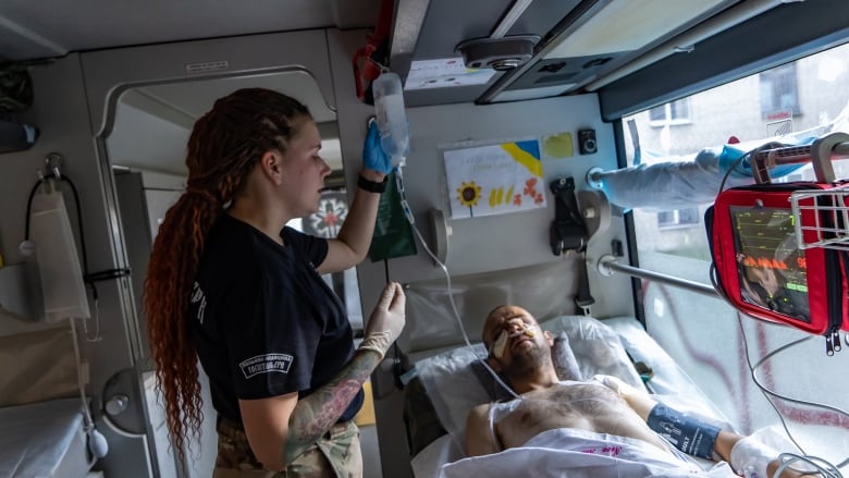 A woman with red braided hair holds a saline drip bag above an injured man in a hospital bed aboard a medical evacuation bus.