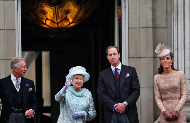 Four people stand on the balcony of a large stone bulding, with large double doors ope  behind them.