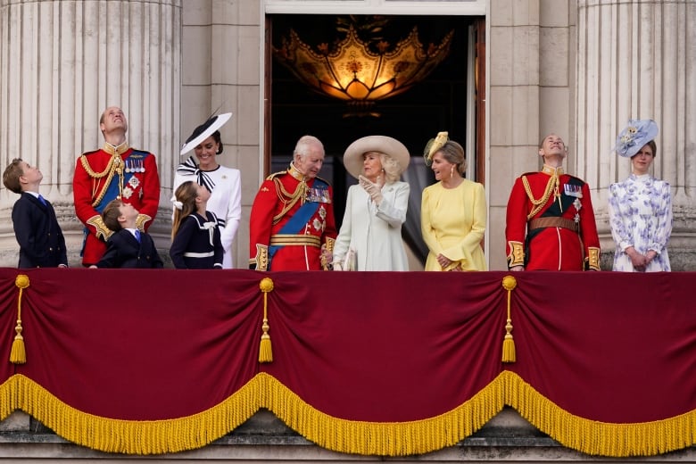 Three children and seven adults stand on the balcony of a large stone building, in front of large open doors. Decorative cloth hangs from the front of the balcony.