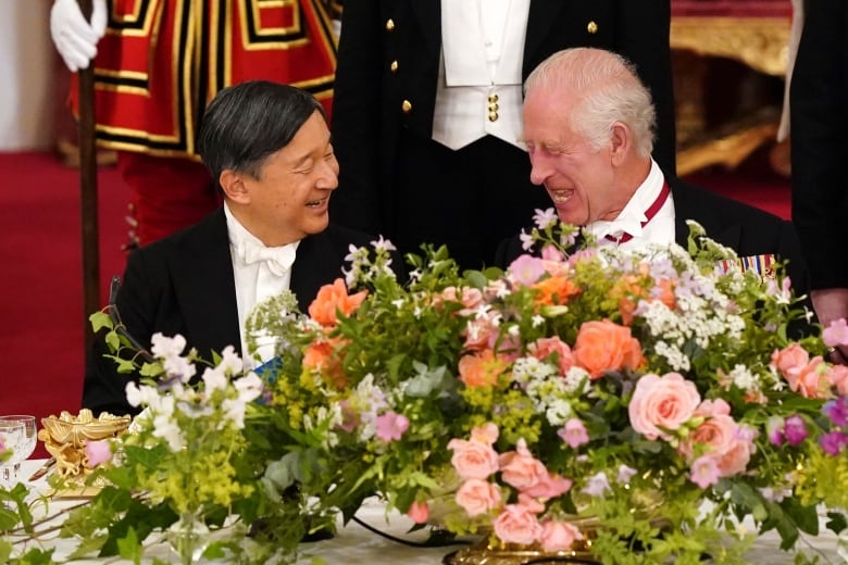 Two people seated at a formal dining table behind a large floral arrangement talk and laugh with one another.