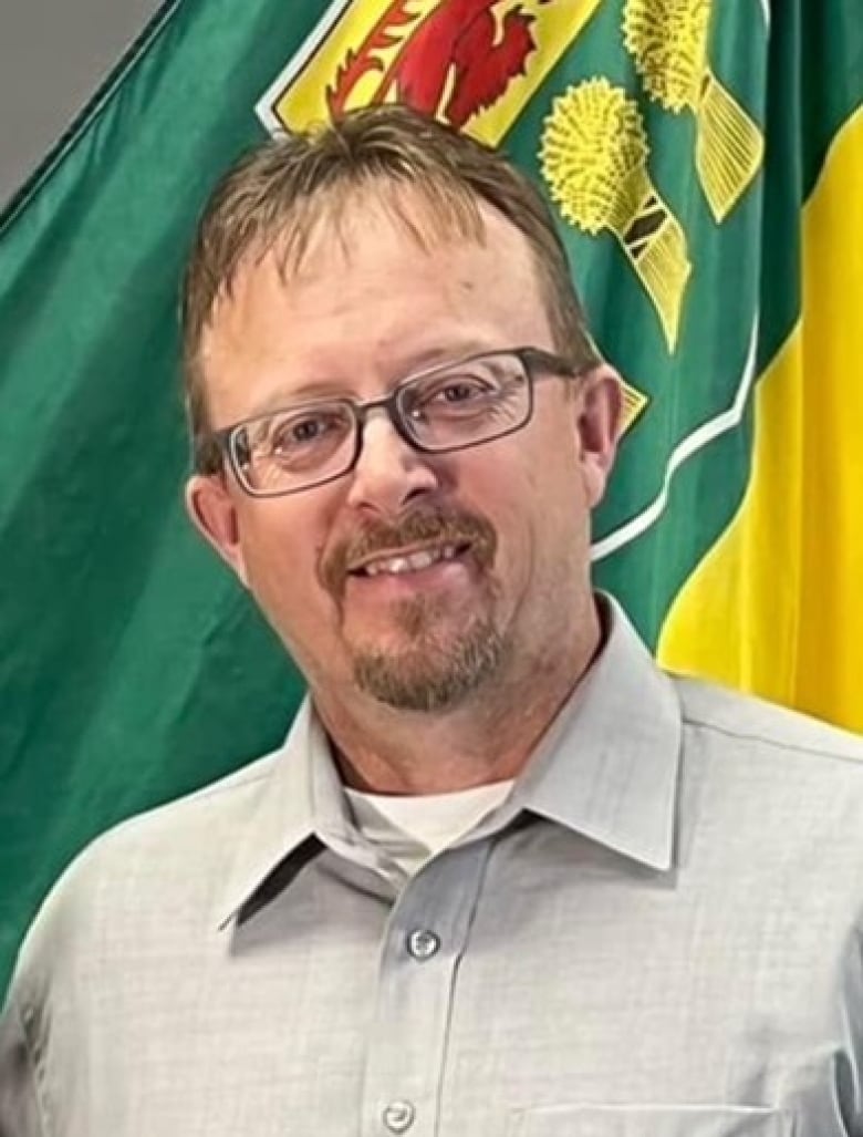 A man with classes stands in front of a furled Saskatchewan flag.