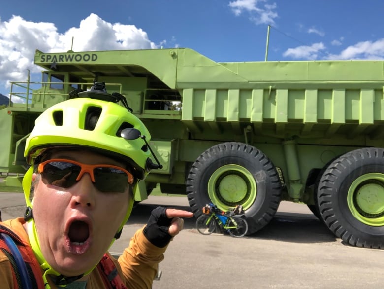 A woman has a shocked expression as she points to a tiny bike beside a huge dump truck. Both are lime green. 