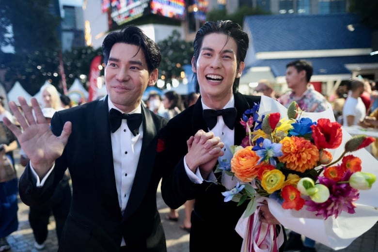 Two people in tuxedos hold hands and gesture during a parade.