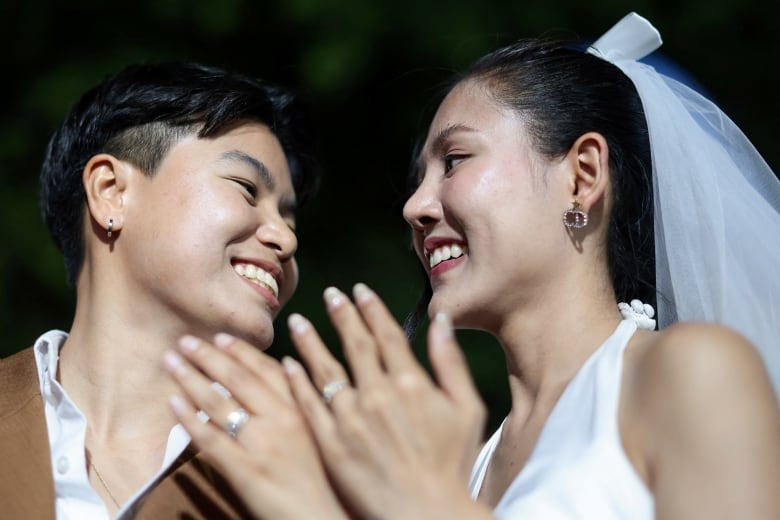 Two people smile at each other as they show off their wedding rings to the camera.