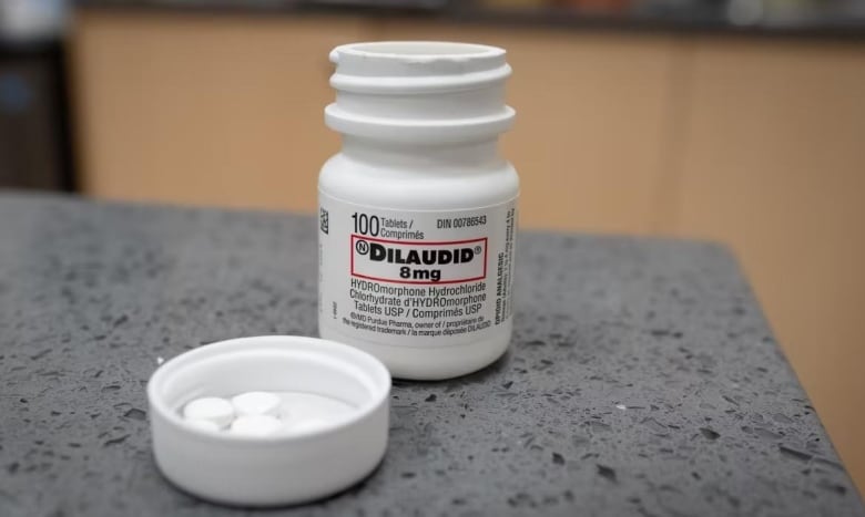 A bottle of white pills sits on a counter with its lid off, and pills in the lid. It reads Dilaudid, 8 mg.