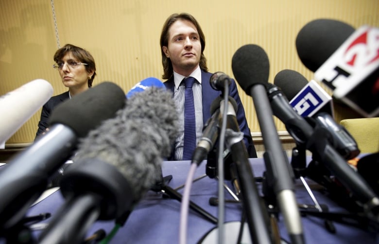 A cleanshaven man in a suit and tie is shown before a bank of microphones.