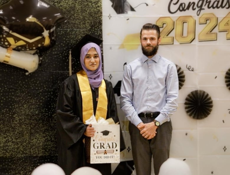 A young woman in a black graduation gown stands next to a bearded man with a collared shirt, standing with his hands clasped.