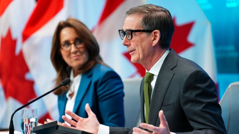 A man and a woman in suits sit behind a desk with microphones, flanked by Canadian flags. 