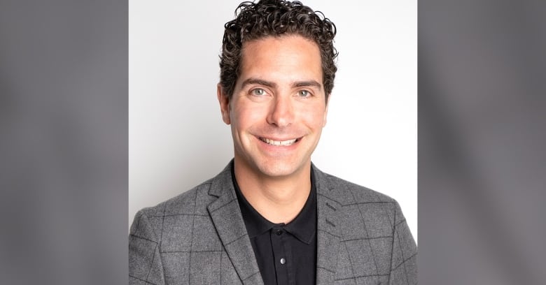 A smiling man in a suit stands in front of a white wall.